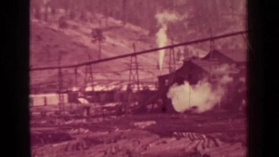 1937 Klamath-area lumber mill seen across a yard, with steam drifting near buildings and wooded hillside behind.