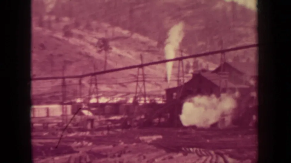 Reddish 8mm view of the Klamath lumber mill yard with a strong steam plume, cable line, small building, and timber piles.