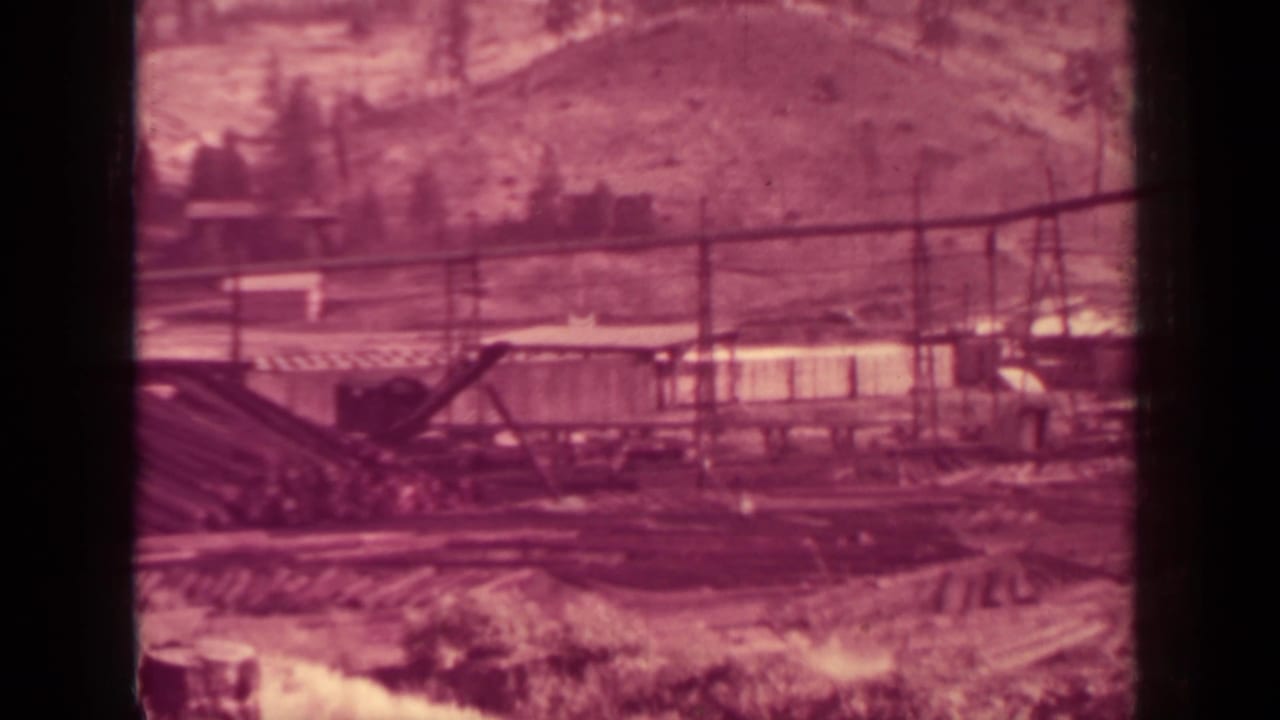 Distant mill yard with crane boom, stacked timber, rail lines, and hillside beyond.