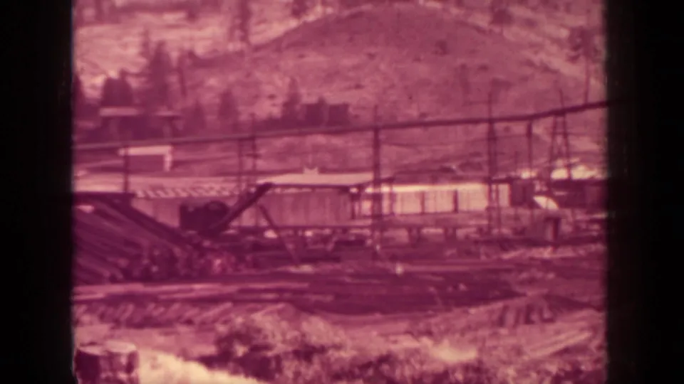 1937 Klamath, California home-movie view of a lumber yard area with crane boom, rail lines, stacked timber, and a hillside backdrop.