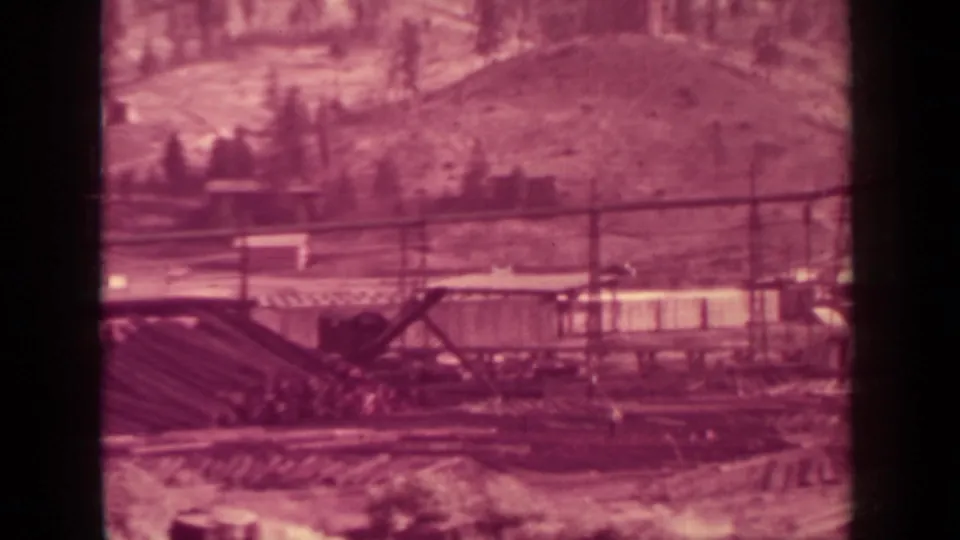Klamath-area 1937 8mm frame showing a crane boom over stacked logs or lumber, rail tracks, poles, and hillside terrain.