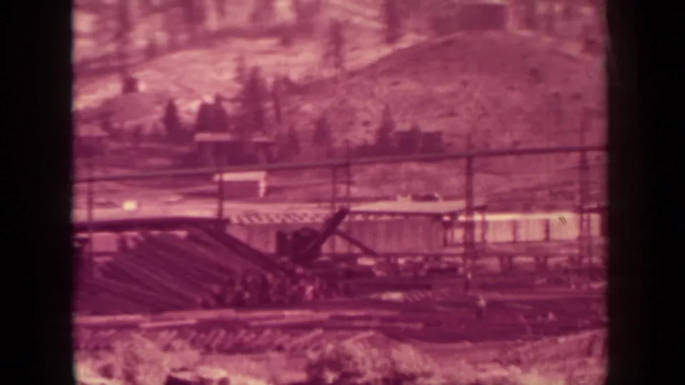 Aged 8mm view from Klamath, California, with timber piles, rail lines, a crane-like structure, low buildings, and a rural hillside.