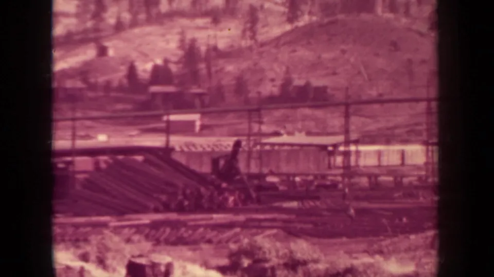 1937 Klamath lumber-yard frame showing rail lines, stacked timber, crane equipment, utility poles, and hillside through reddish-purple aged film.