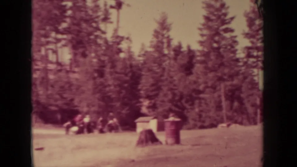 Klamath-area forest clearing with distant people gathered near trees, plus a stump and red barrel in the foreground.