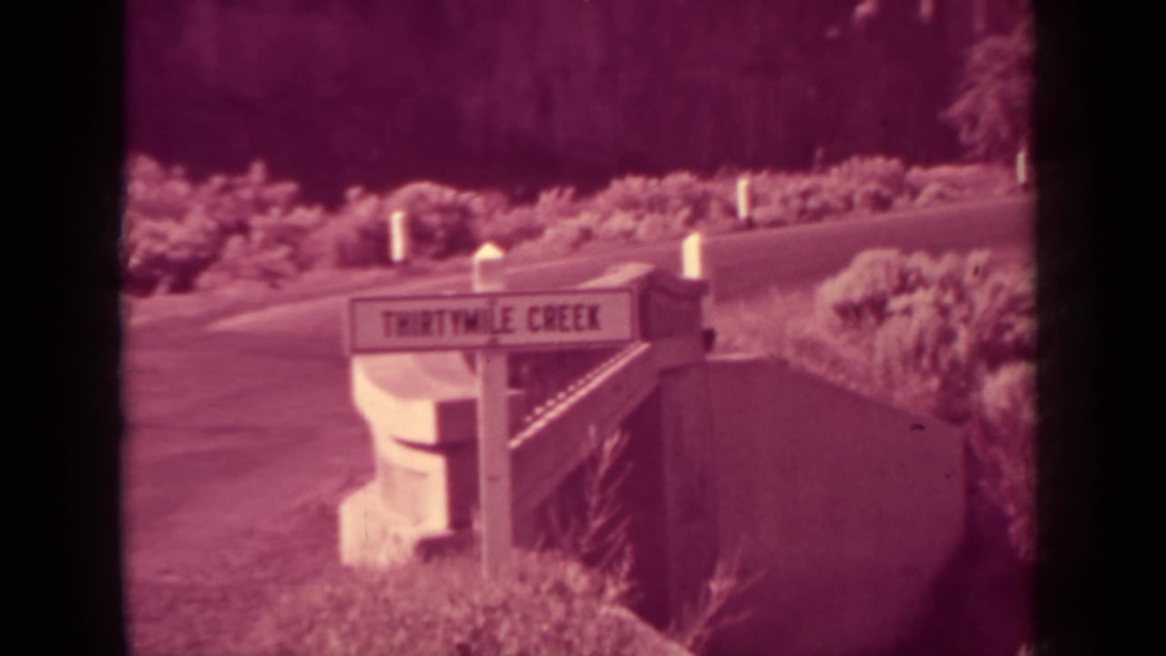 Thirtymile Creek sign beside a winding road with brush and hillside in the background.