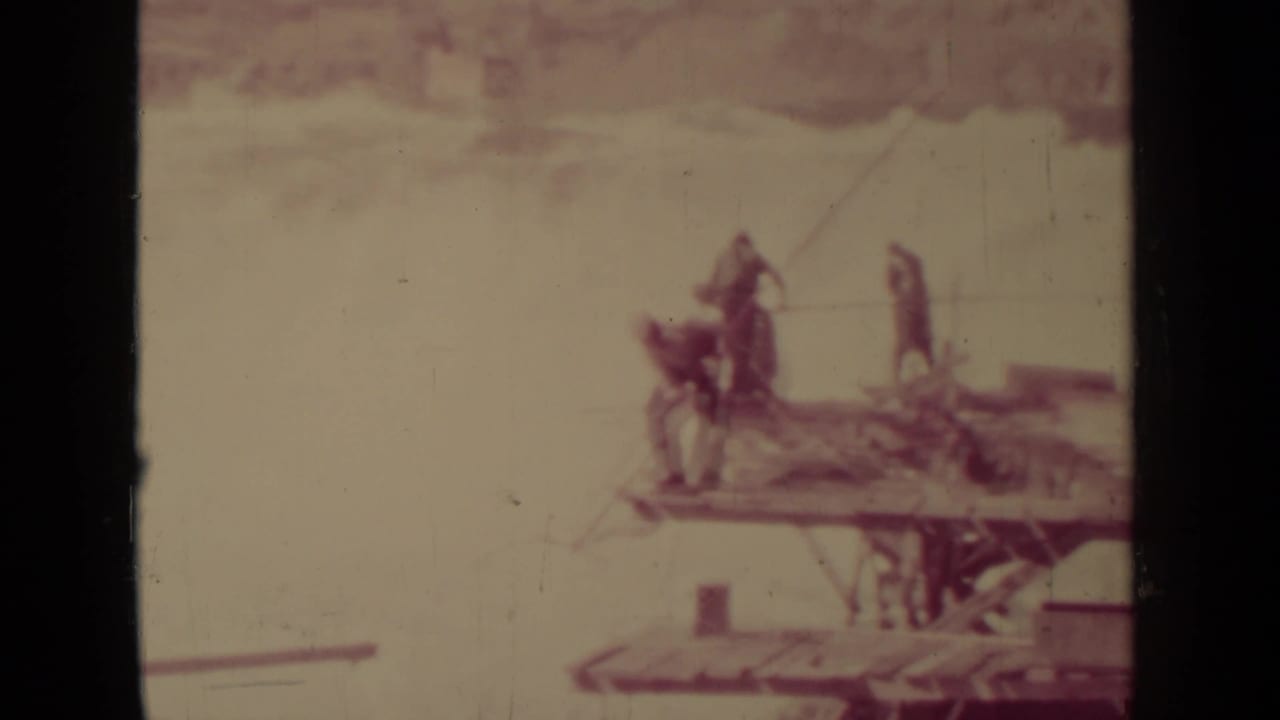 Small figures stand on a wooden platform near rushing water, partly obscured by film gate shadows.