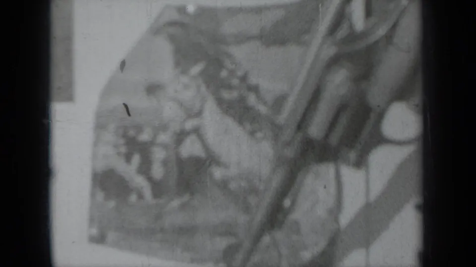 Black-and-white frame from Klamath, California, showing a long gun handled near a blurred rodeo poster.