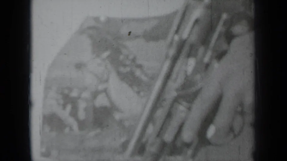 A diagonal long gun and partial hands appear in front of a rodeo-themed poster in black-and-white 8mm footage.