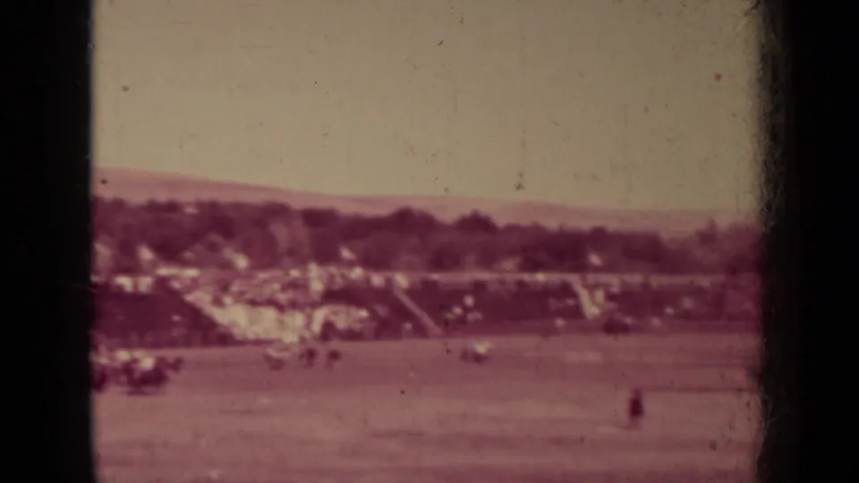 1937 Klamath, California 8mm frame showing a reddish-tinted baseball field with a grandstand, distant spectators, trees, and low hills.