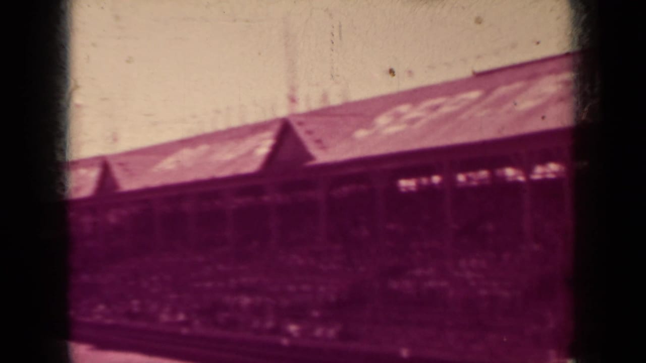 A long Klamath ballpark grandstand fills the frame, with roof lettering and a shaded crowd visible under the structure.