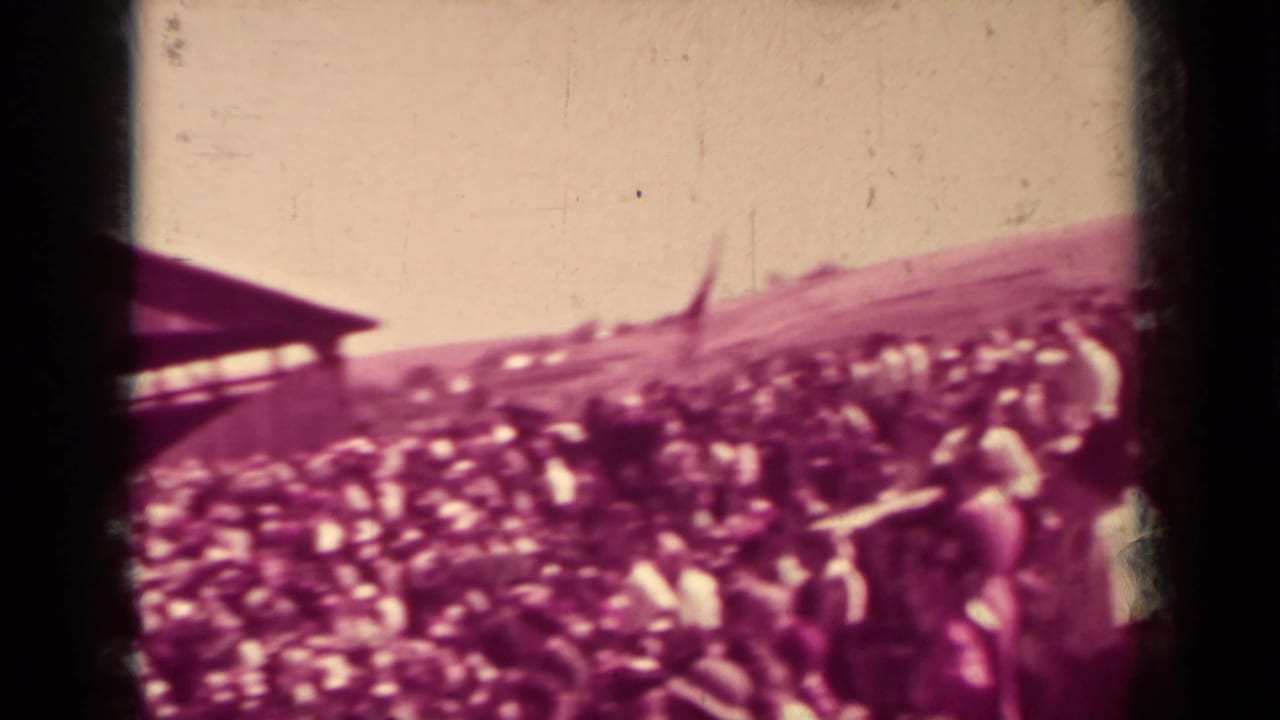 Crowded outdoor gathering seen across a sloped open area with dark film-frame edges.