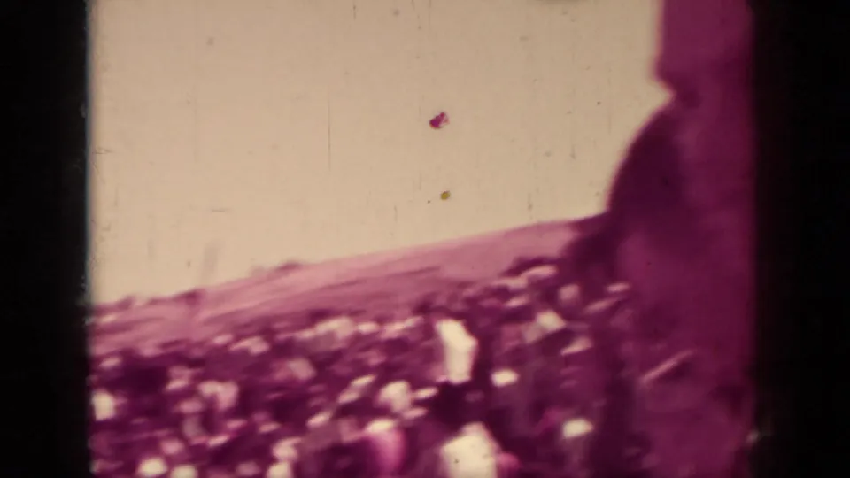 Handheld 8mm view from within a Klamath public gathering, looking past a standing figure toward a dense outdoor crowd.
