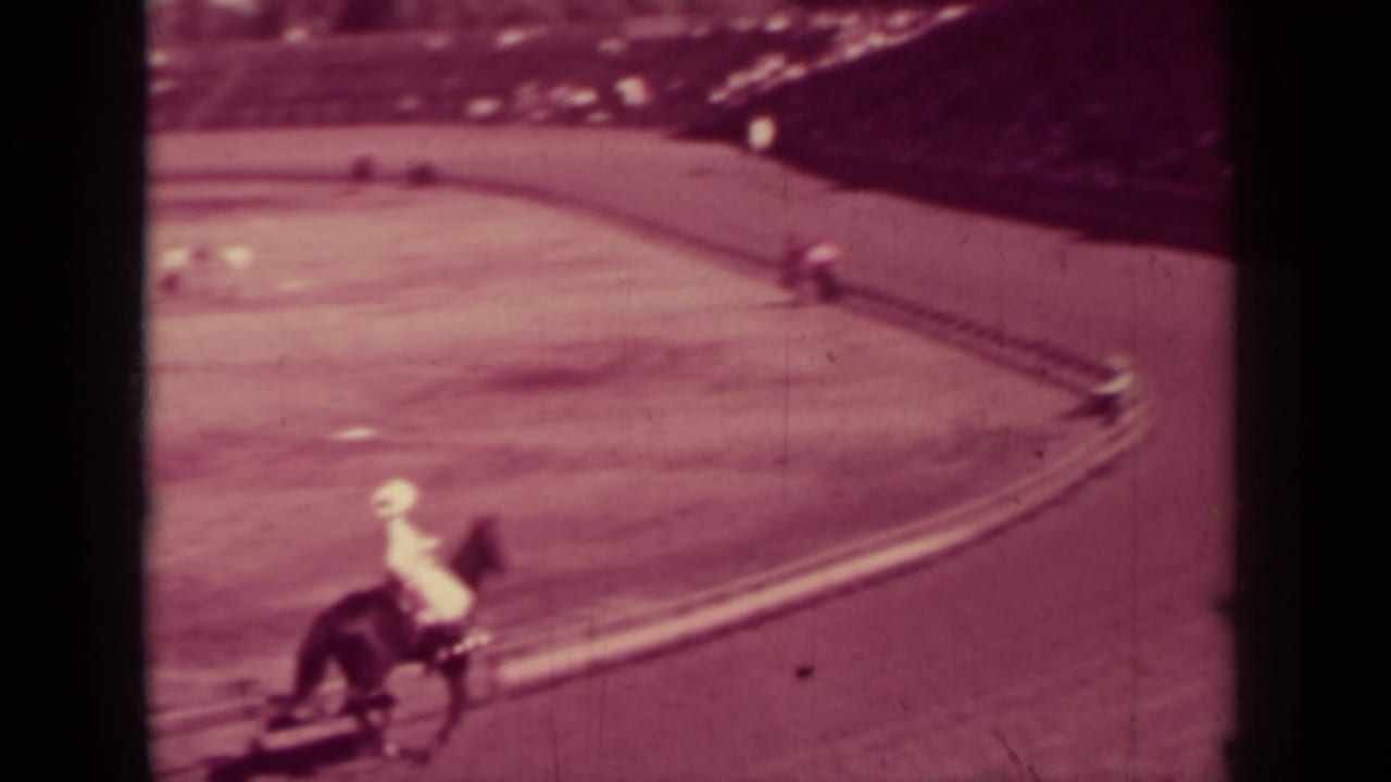 Mounted rider rounding a dirt racetrack bend near a rail with spectators beyond.