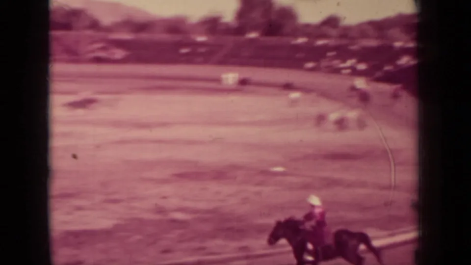 Soft 8mm frame of a rider on a dark horse moving along a dusty racetrack, with distant riders and spectators in the background.