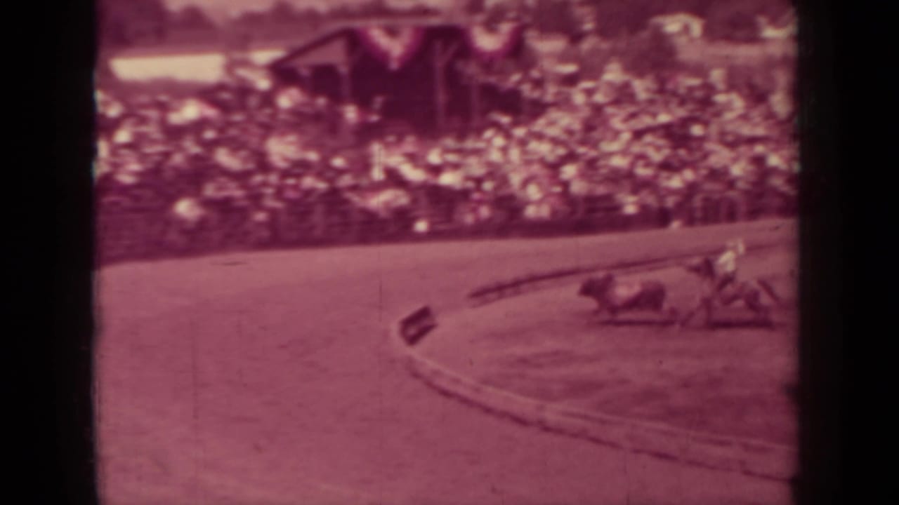 Horses and riders round a dirt track with crowded stands in the background.