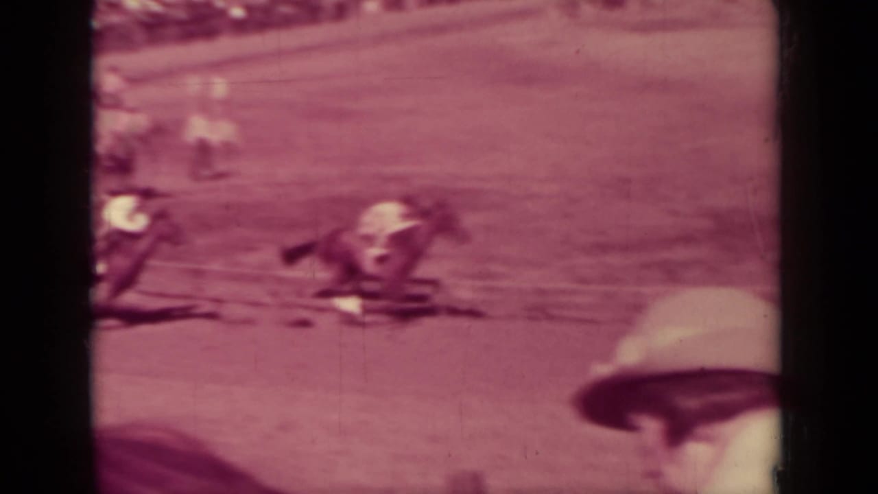 Close pass of a horse and rider on a 1937 Klamath dirt track, partly obscured by a foreground spectator hat or helmet.