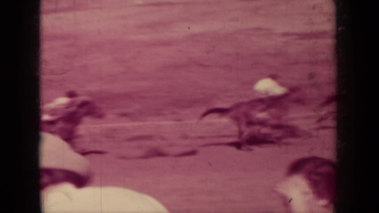 Horses and jockeys race past spectators at the edge of a dusty track.