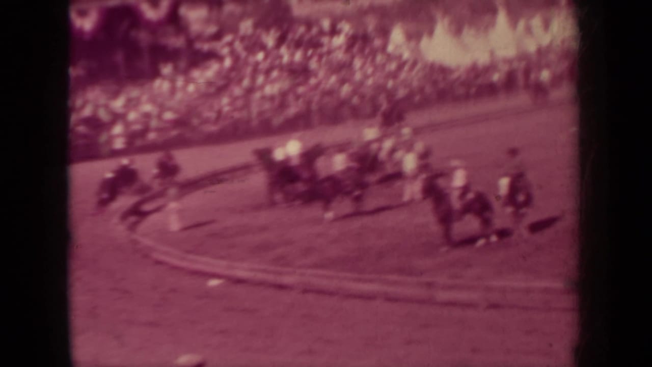 Horses and riders move around a dirt track before a crowded stand.