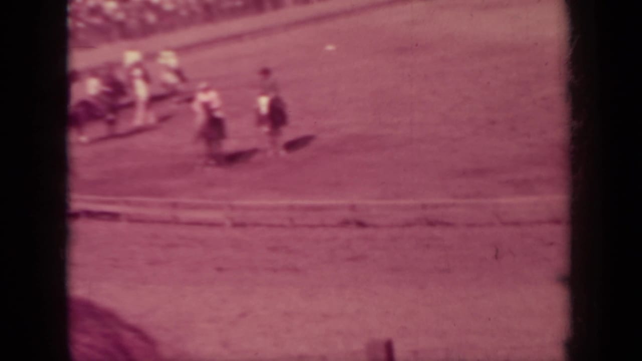 Horses and riders appear on the far curve of a rural fairground dirt track, with the infield and rail visible in soft 8mm focus.