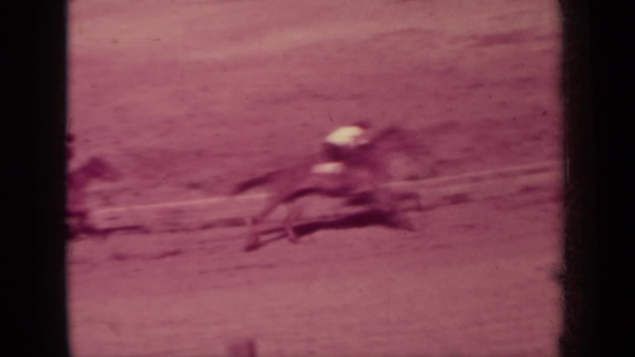 A horse and rider rush past the camera position in a motion-blurred 8mm frame, emphasizing the speed of the fairground race.