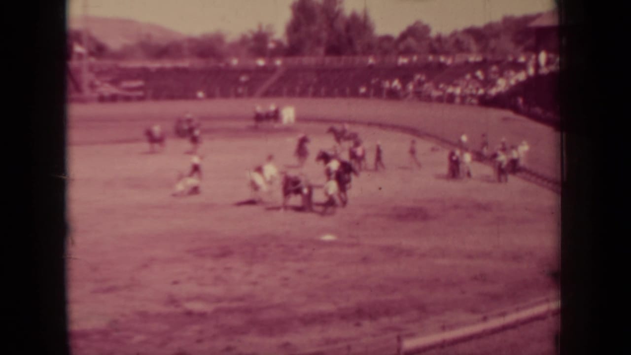 Wide 1937 Klamath fairground view with riders on the dirt oval, spectators in the stands, trees beyond, and the infield visible.