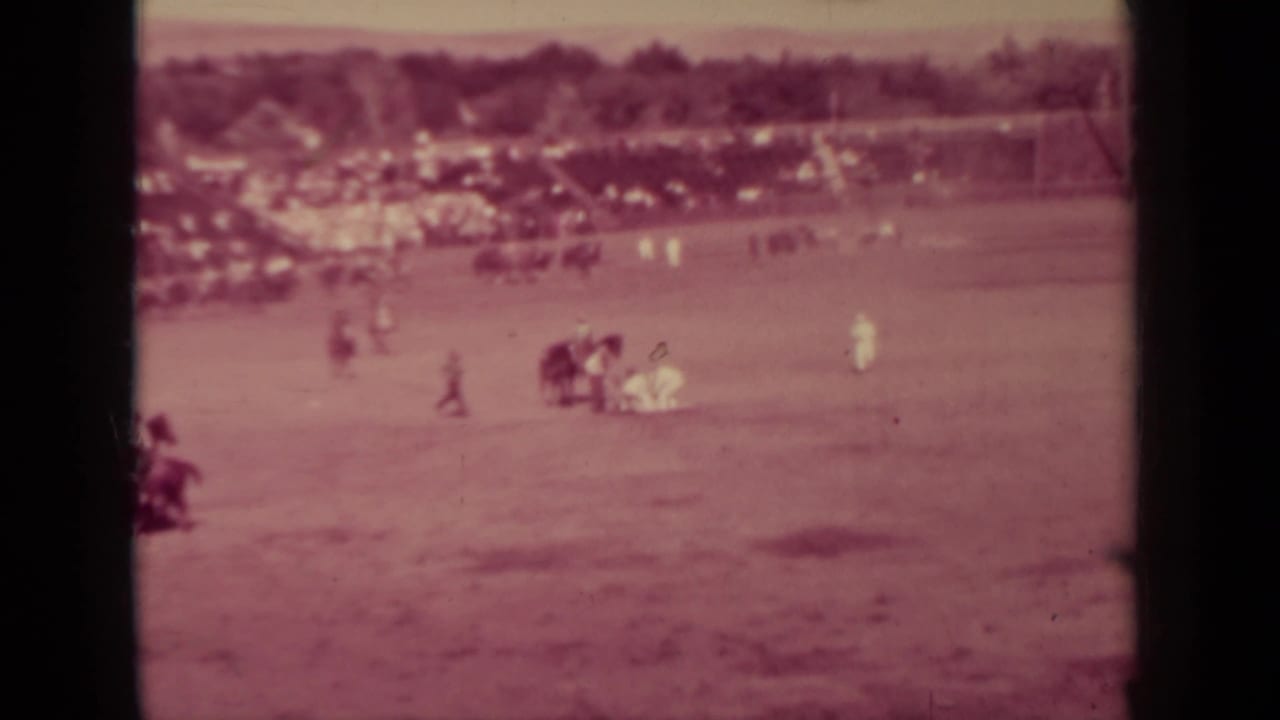 Distant riders and people spread across a dusty field with bleachers in the background.