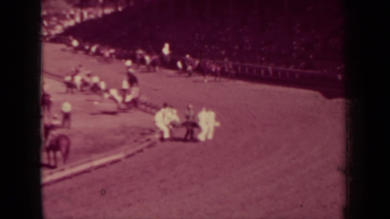 Dirt racetrack with attendants gathered near a fallen jockey as spectators sit in the grandstand.
