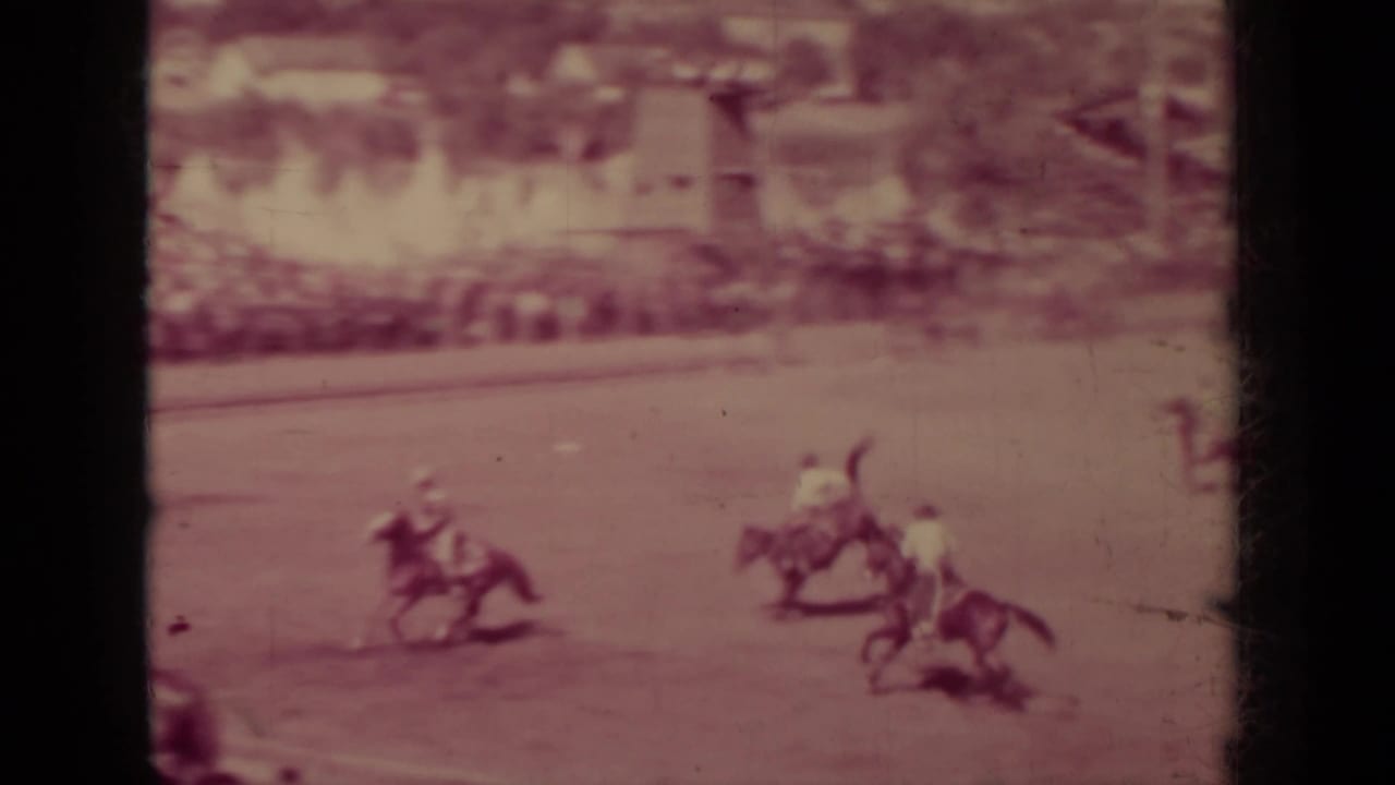 Riders on horseback cross a dirt rodeo arena with distant spectators in the background.