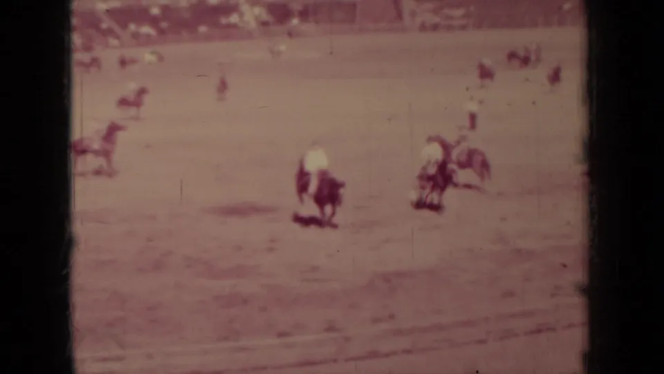 Soft 8mm frame of a rider in the Klamath rodeo arena, with additional mounted figures and spectators beyond the field.