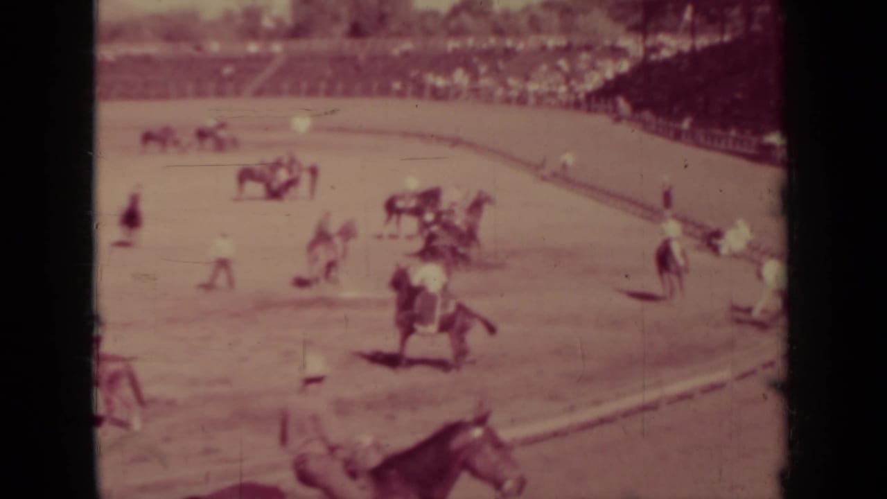 Jockeys ride horses around a dirt track with spectators in the background.