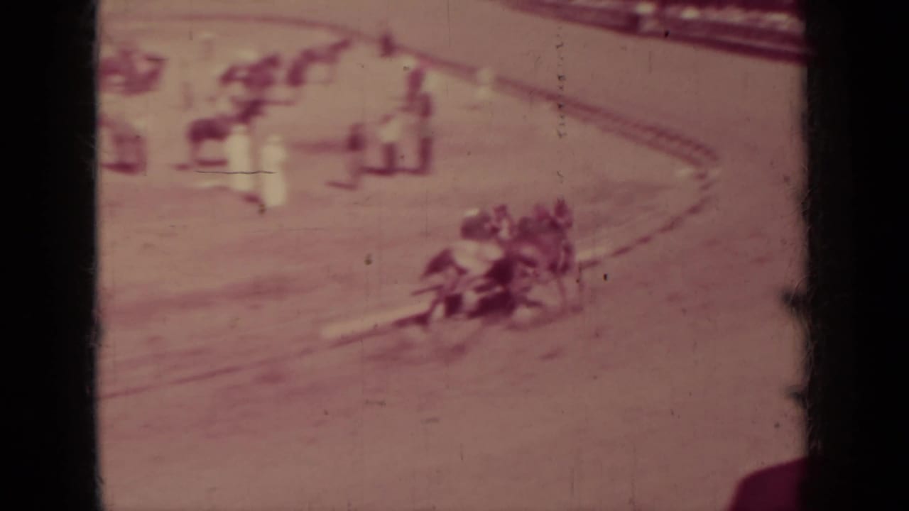 Jockeys ride horses around a dirt track with blurred stands in the background.