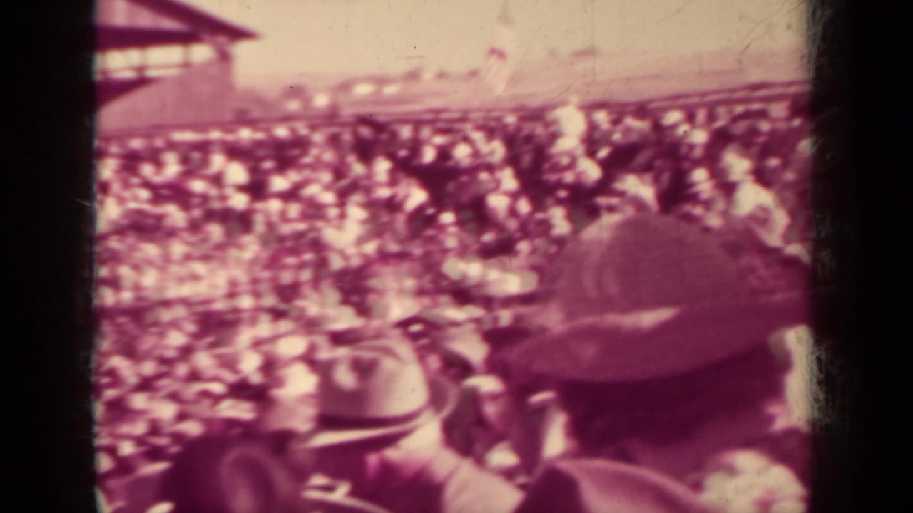 Crowded spectator area with hats in the foreground and a pavilion audience beyond.