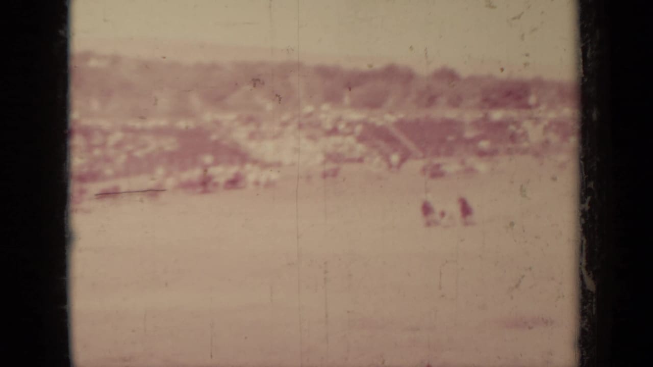 Faded 8mm view of Klamath, California with open ground, distant buildings, and small human figures barely visible near the background.