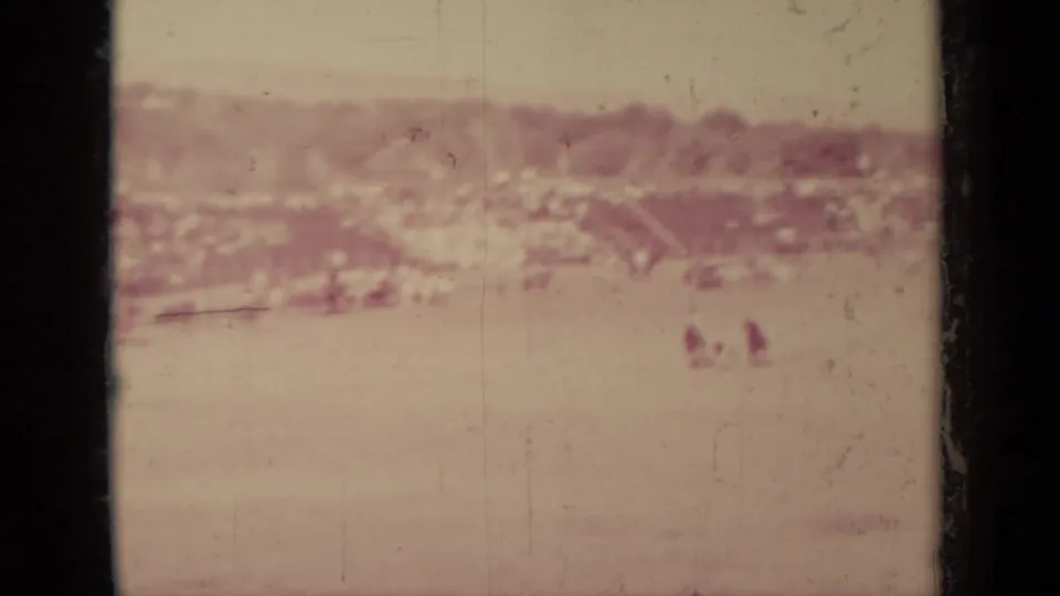 Faded 8mm view of Klamath, California with open ground, distant buildings, and small human figures barely visible near the background.