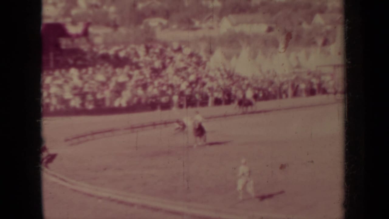 Rodeo arena curve with rider, livestock, crowd, fencing, and a small building beyond the stands.