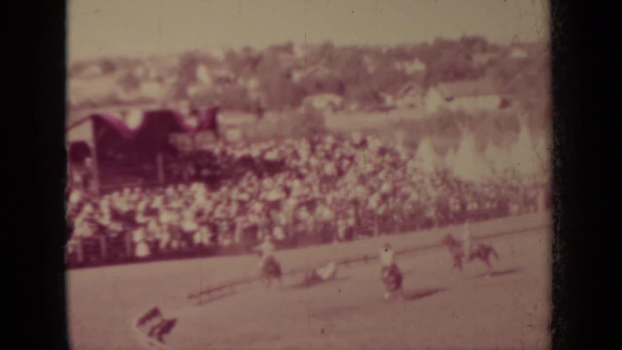 Wide home-movie view of the Klamath rodeo crowd and arena, with riders and livestock visible near the fence line.