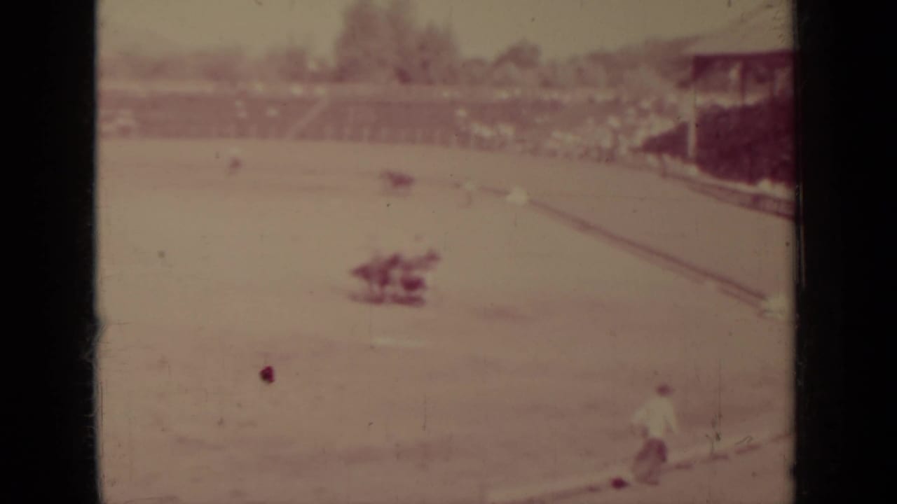 A rider crosses the open dirt track with spectators and arena structures in the background.