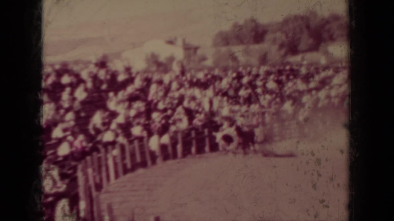 Wide rodeo arena with bleachers of spectators behind a rail fence.