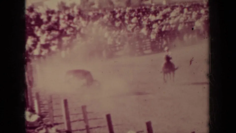 Wide 8mm frame from the Klamath rodeo showing a rider on horseback and livestock in a dusty fenced arena with spectators beyond.