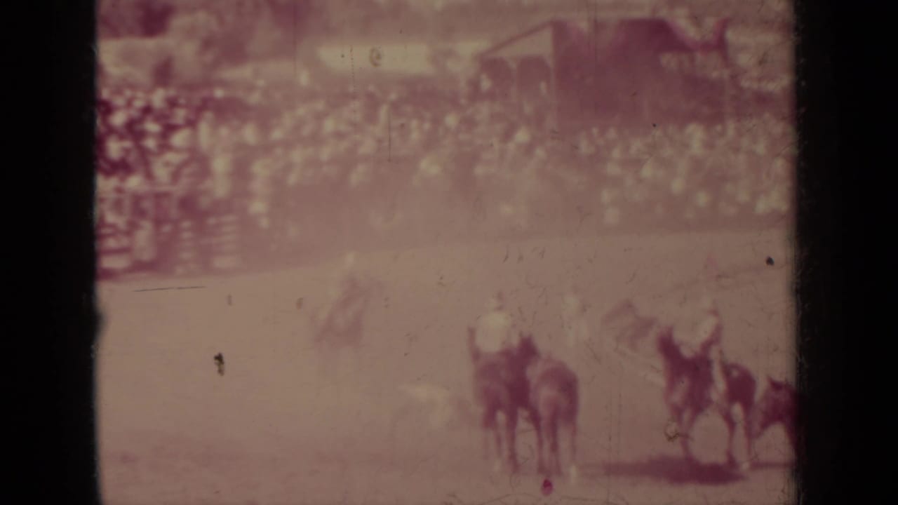 Mounted cowboys gather or pass in the Klamath rodeo arena, with bleachers and a wooden structure behind them.