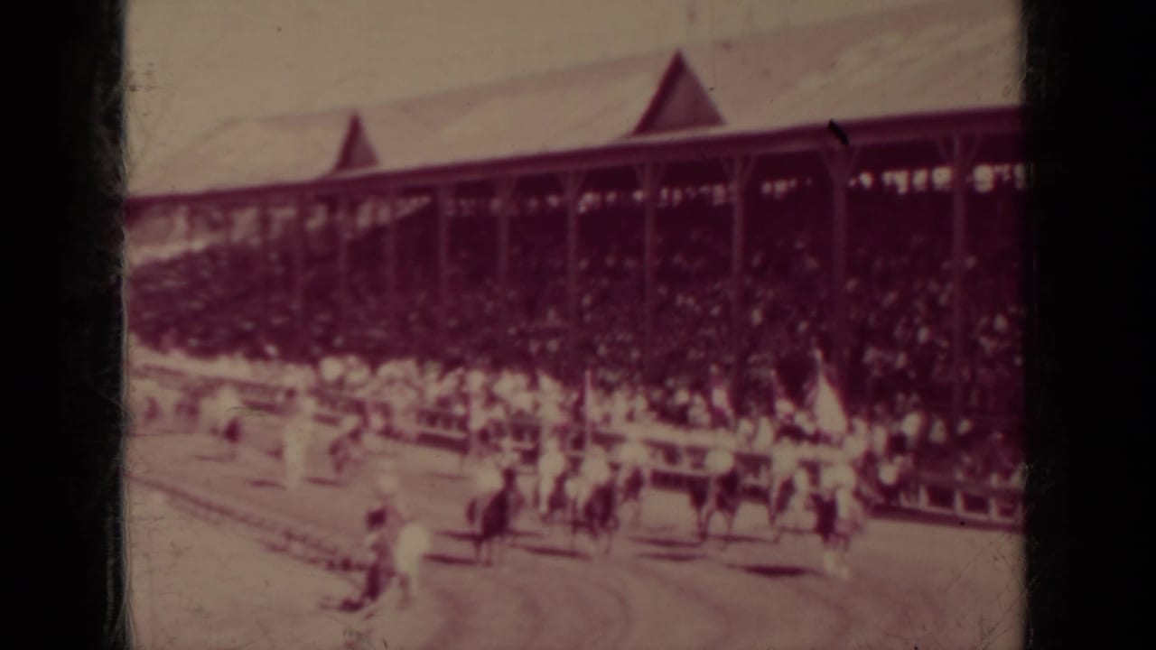 Horses and riders pass in front of a crowded covered grandstand.