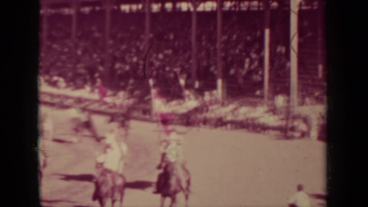 Mounted riders pass an arena crowd behind railings.