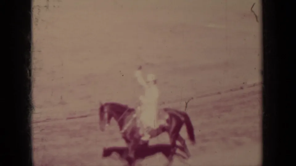 A mounted rider in light-colored clothing raises an arm in greeting or signal within the open arena setting.