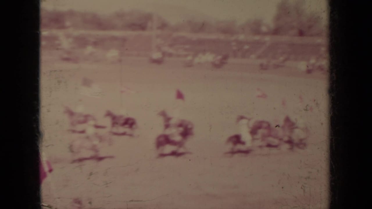 Distant horseback riders move across an outdoor arena with spectators beyond the field.