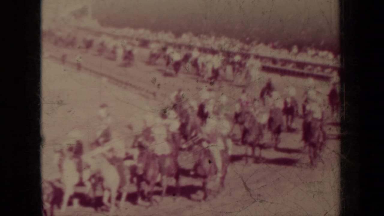 Cluster of mounted riders near a rail line with spectators gathered behind them.