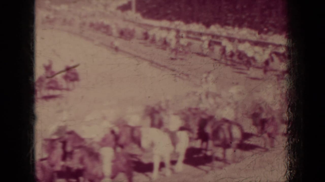 Mounted riders and horses pass through a dusty outdoor area with spectators in the background.