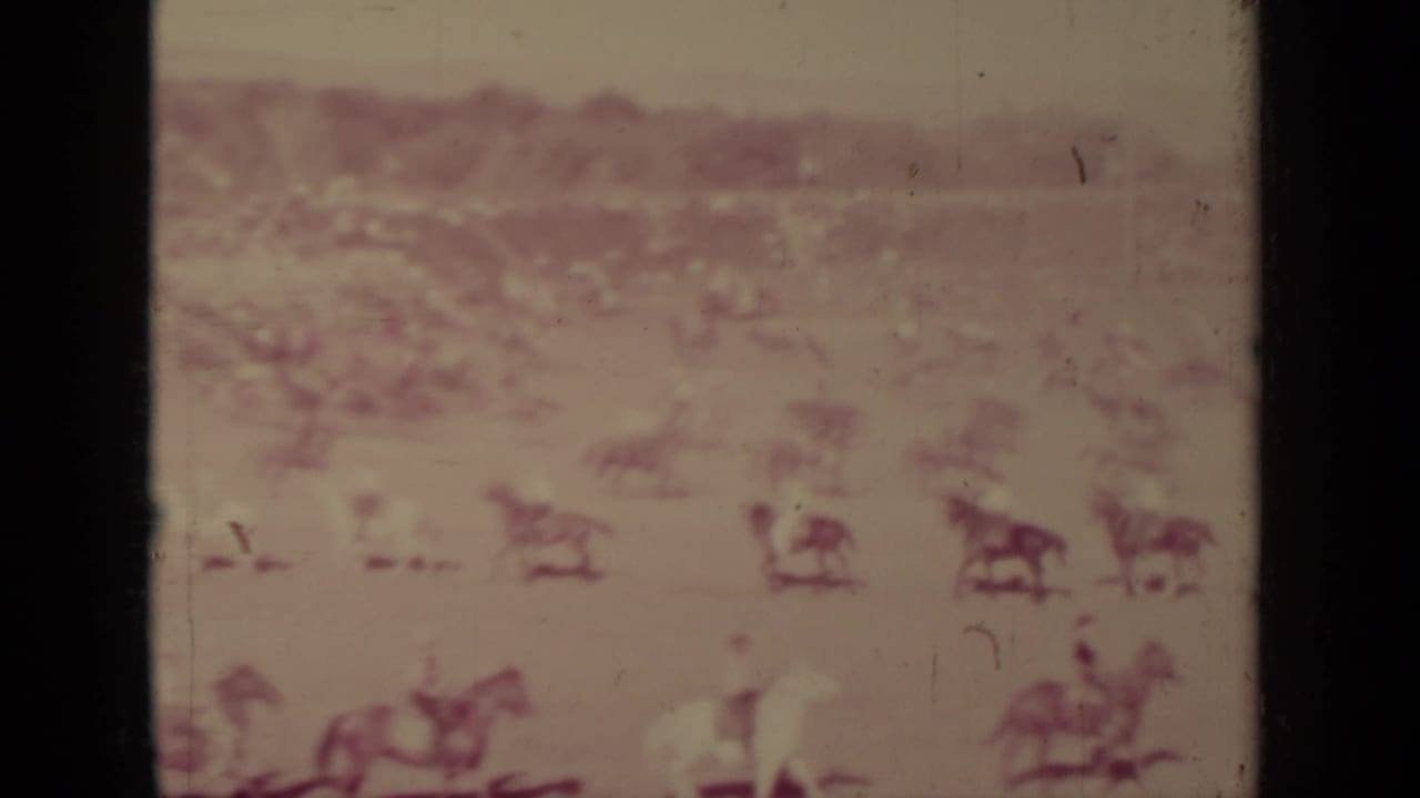 Distant horseback riders spread across a dusty arena with dark film borders.