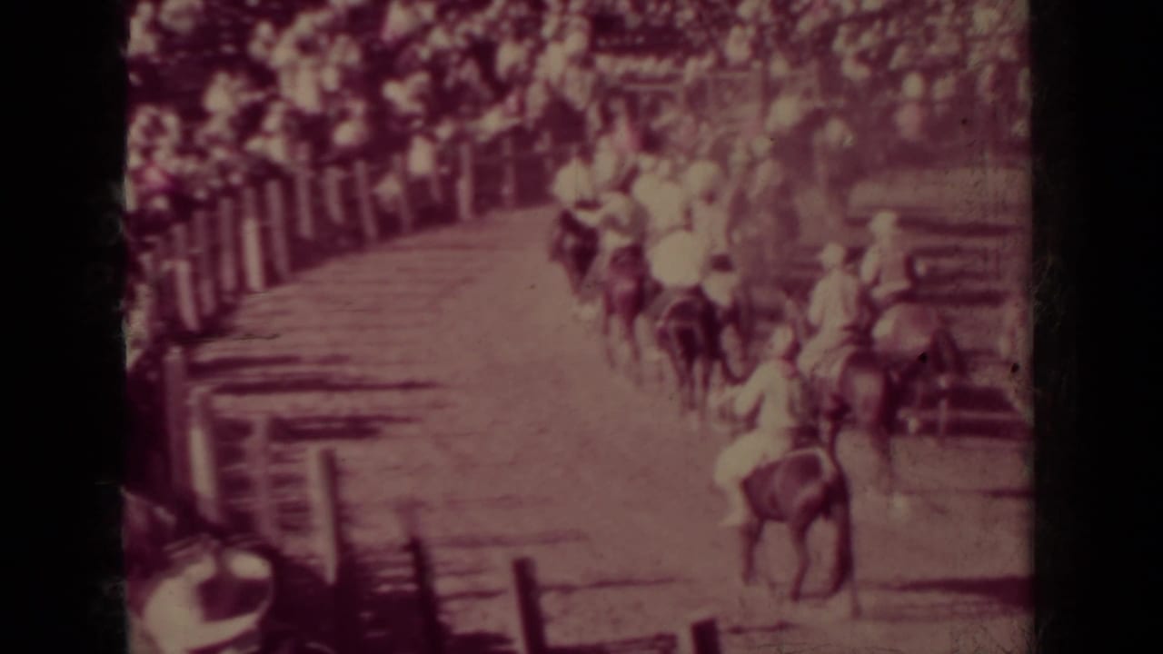 Mounted riders move along a dirt track beside a wooden rail and shaded spectators.