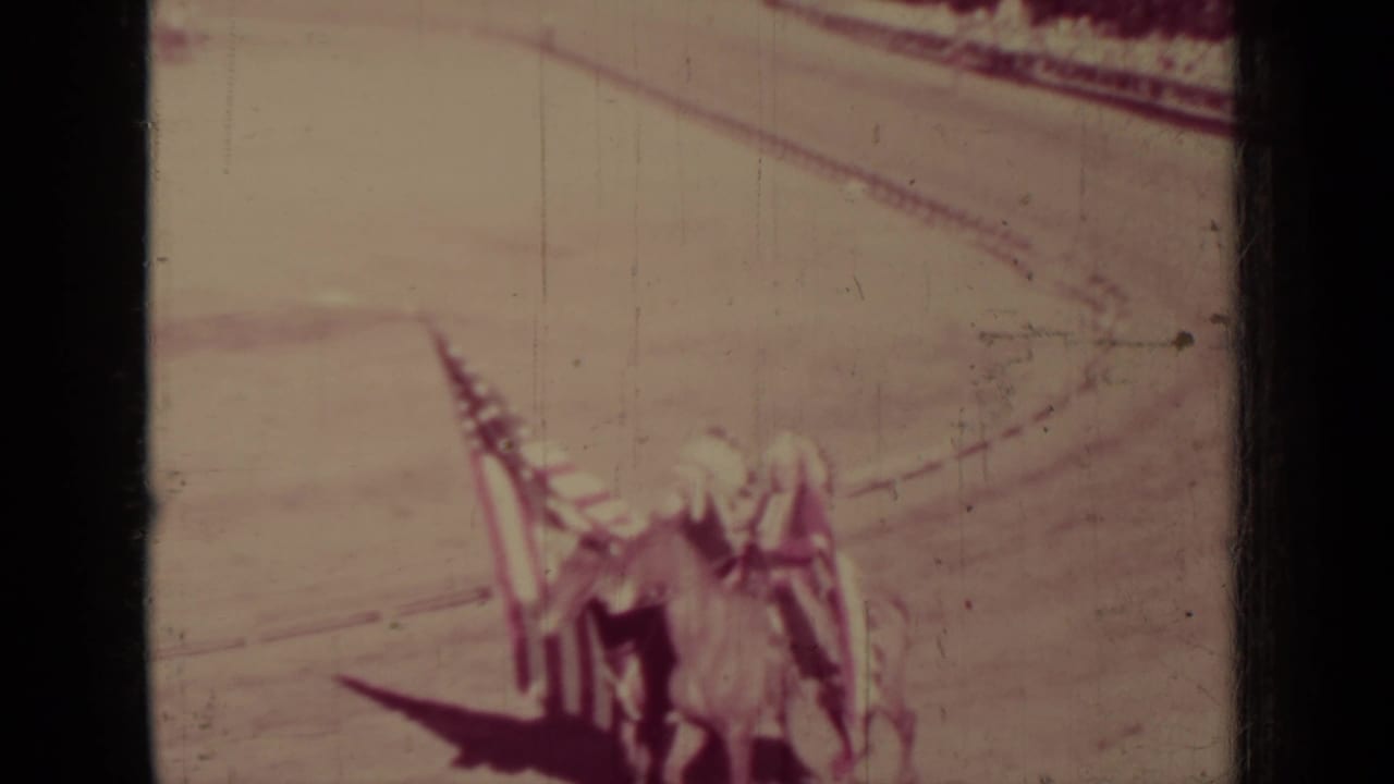 Distant horseback riders in costume cross a sandy arena with a flag visible above them.