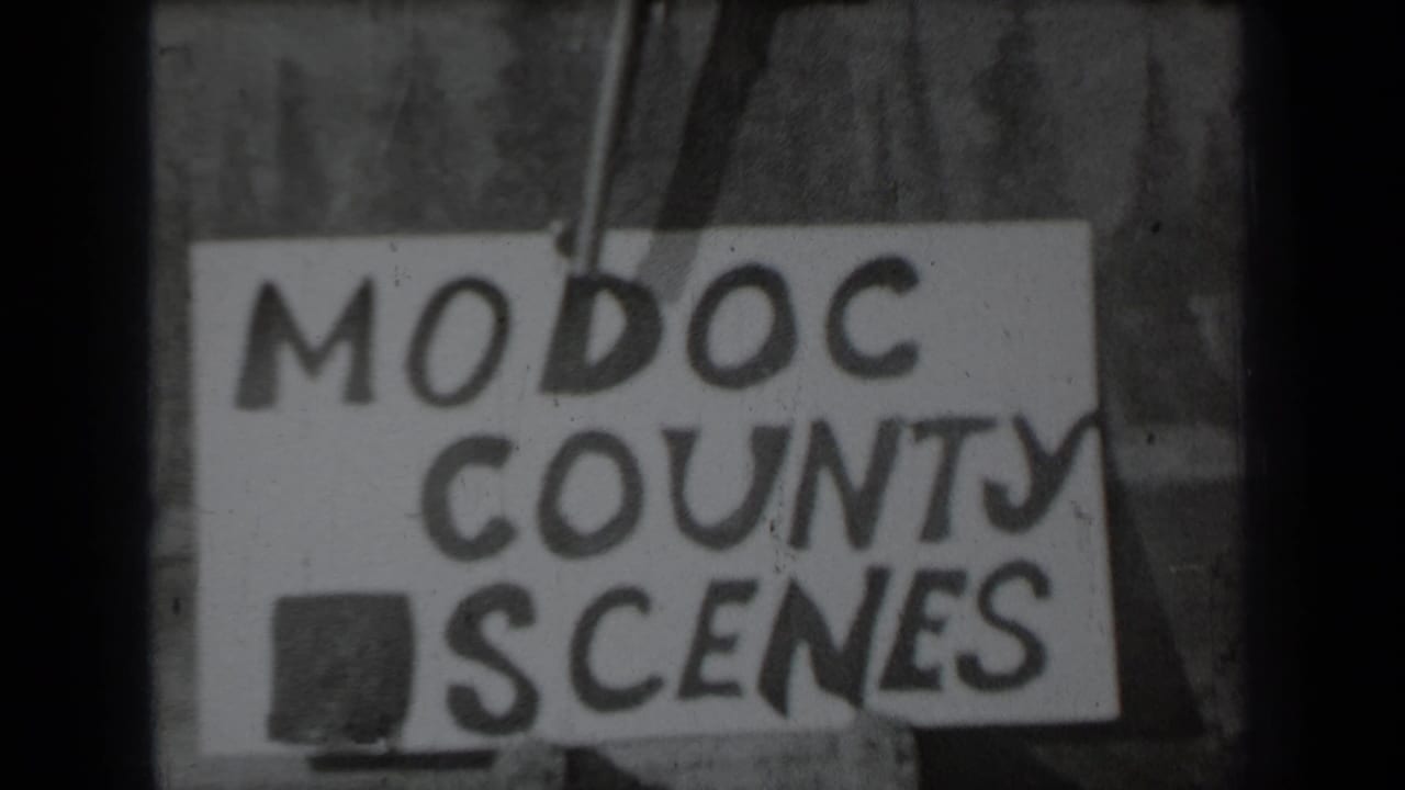 Black-and-white 1937 8mm frame showing a hand holding a revolver above a hand-lettered Modoc County Scenes title card.